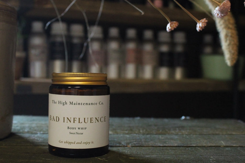 A whipped body butter jar from The High Maintenance Co. named 'Bad Influence', featuring a dark amber glass bottle, a gold metal lid, and a cream-colored label, set on a rustic wooden table with dried flowers in the background.