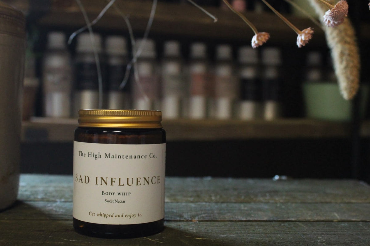 A whipped body butter jar from The High Maintenance Co. named 'Bad Influence', featuring a dark amber glass bottle, a gold metal lid, and a cream-colored label, set on a rustic wooden table with dried flowers in the background.