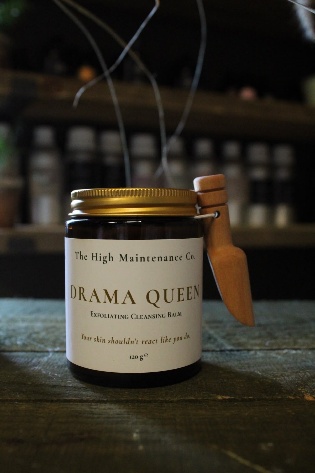 A dark glass jar of 'Drama Queen' Exfoliating Cleansing Balm by The High Maintenance Co., featuring a gold metal lid, a white label, and a wooden spatula tool attached to the side, set on a rustic wooden surface.