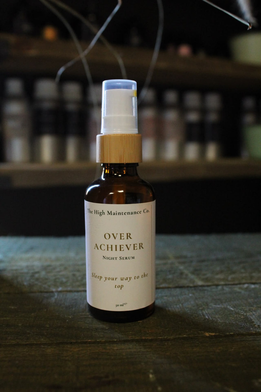 An amber glass pump bottle labelled 'OVER ACHIEVER Night Serum' by The High Maintenance Co, featuring a bamboo-look pump collar and white spray top, set on a dark wooden surface with a blurred background of shelves and bottles. 