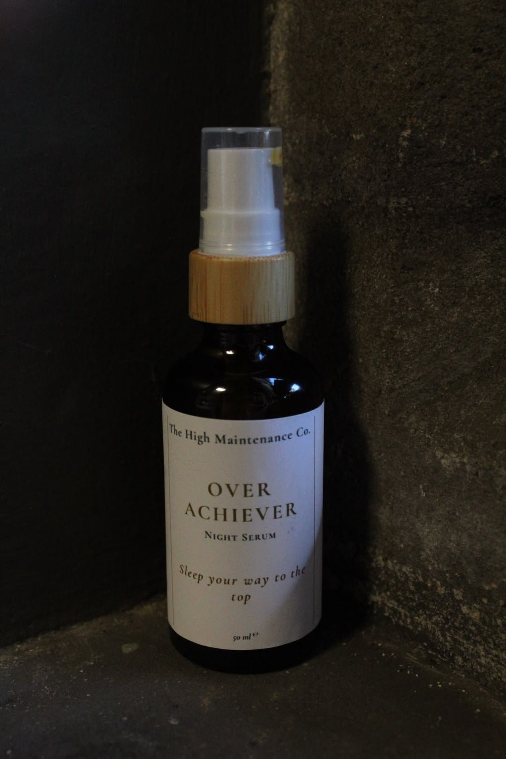 A dark amber glass night serum bottle with a white label and a light wood-toned collar, standing in a dark textured corner. The label reads 'The High Maintenance Co.