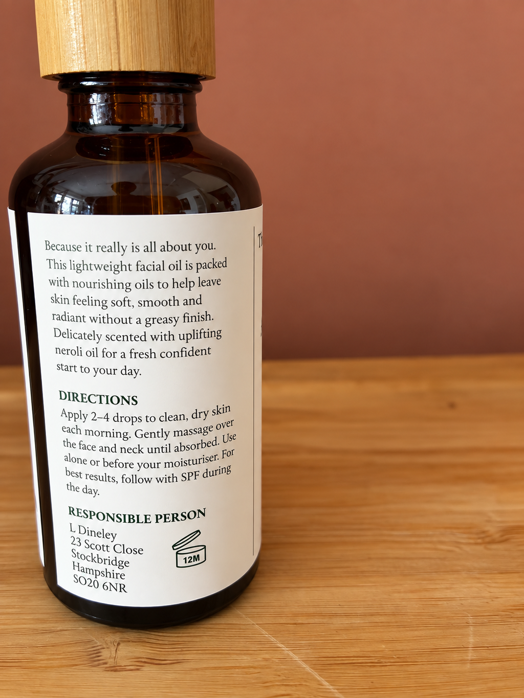 An amber glass bottle of facial oil with a light wood cap, showing the back label with product details, set on a wooden surface against a terracotta background.