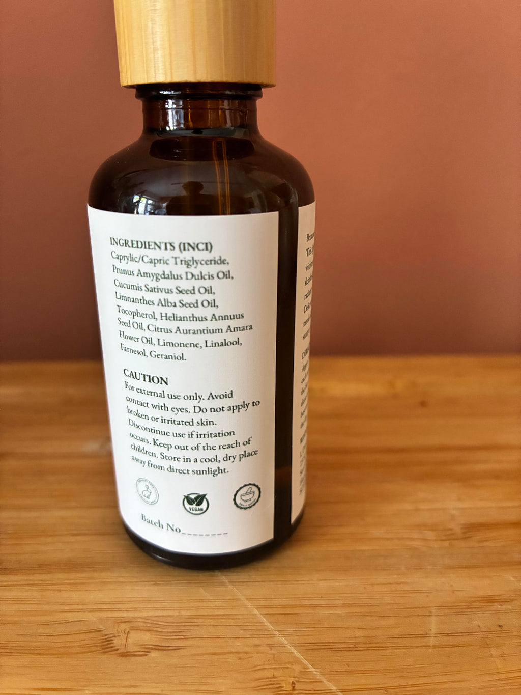 An amber glass bottle of face serum with a light wood cap and a white label, resting on a wooden surface against a terracotta background.