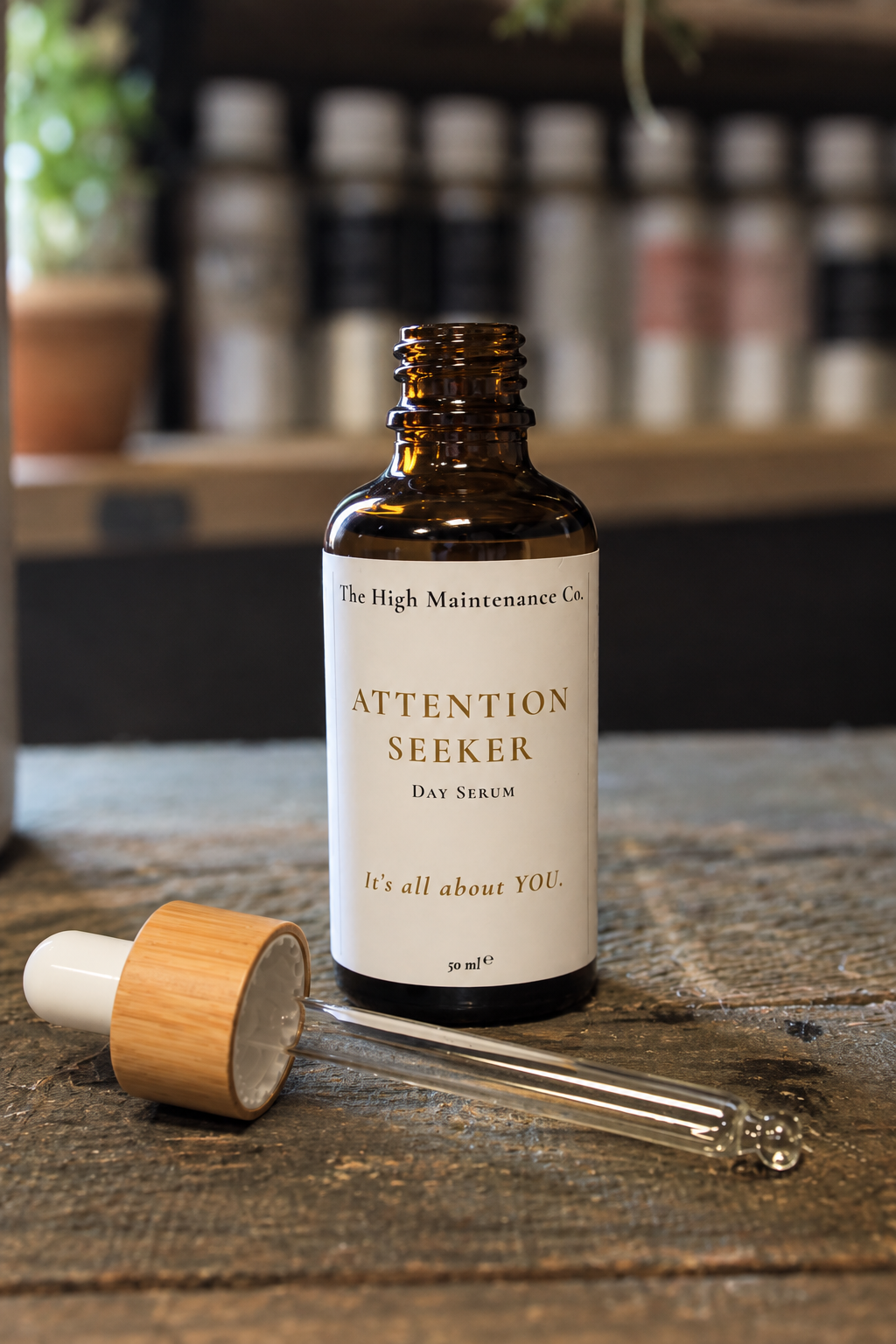 An amber glass dropper bottle labeled 'Attention Seeker Day Serum' by The High Maintenance Co., shown on a rustic wooden surface with its dropper cap beside it and a softly blurred shelf of essential oil bottles in the background.