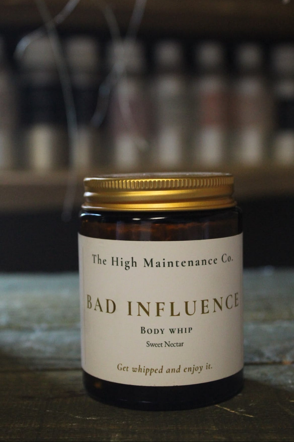 An amber glass jar of organic body whip fragranced with honey and vanilla with a gold metal lid and a cream-colored label reading 'The High Maintenance Co. BAD INFLUENCE Body Whip Sweet Nectar' sits on a dark wooden surface with a blurred shop background.
