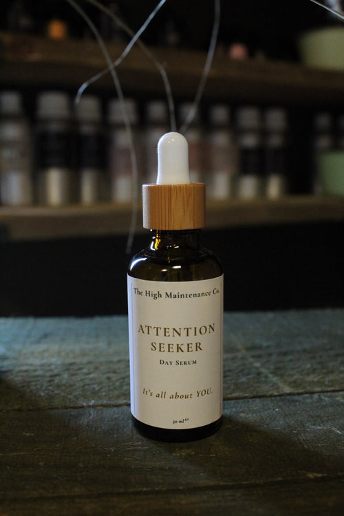 A 50 ml amber glass dropper bottle labeled 'Attention Seeker Day Serum' by The High Maintenance Co., standing on a dark wooden surface with blurred shelves in the background.
