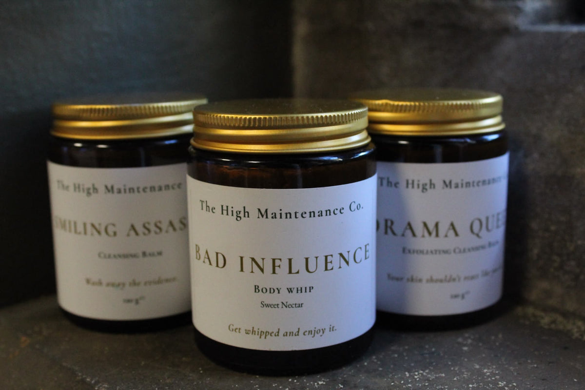 Three amber glass jars of organic skin care products with gold lids and white labels from The High Maintenance Co., arranged on a dark textured surface.