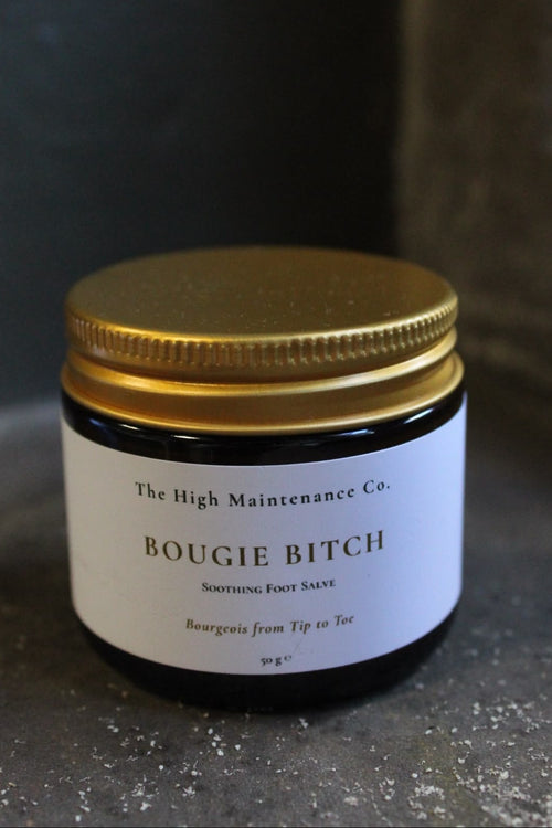 A dark amber glass jar with a metallic gold screw-on lid and a white paper label reading 'The High Maintenance Co. BOUGIE BITCH Soothing Foot Salve' on a dark textured surface.