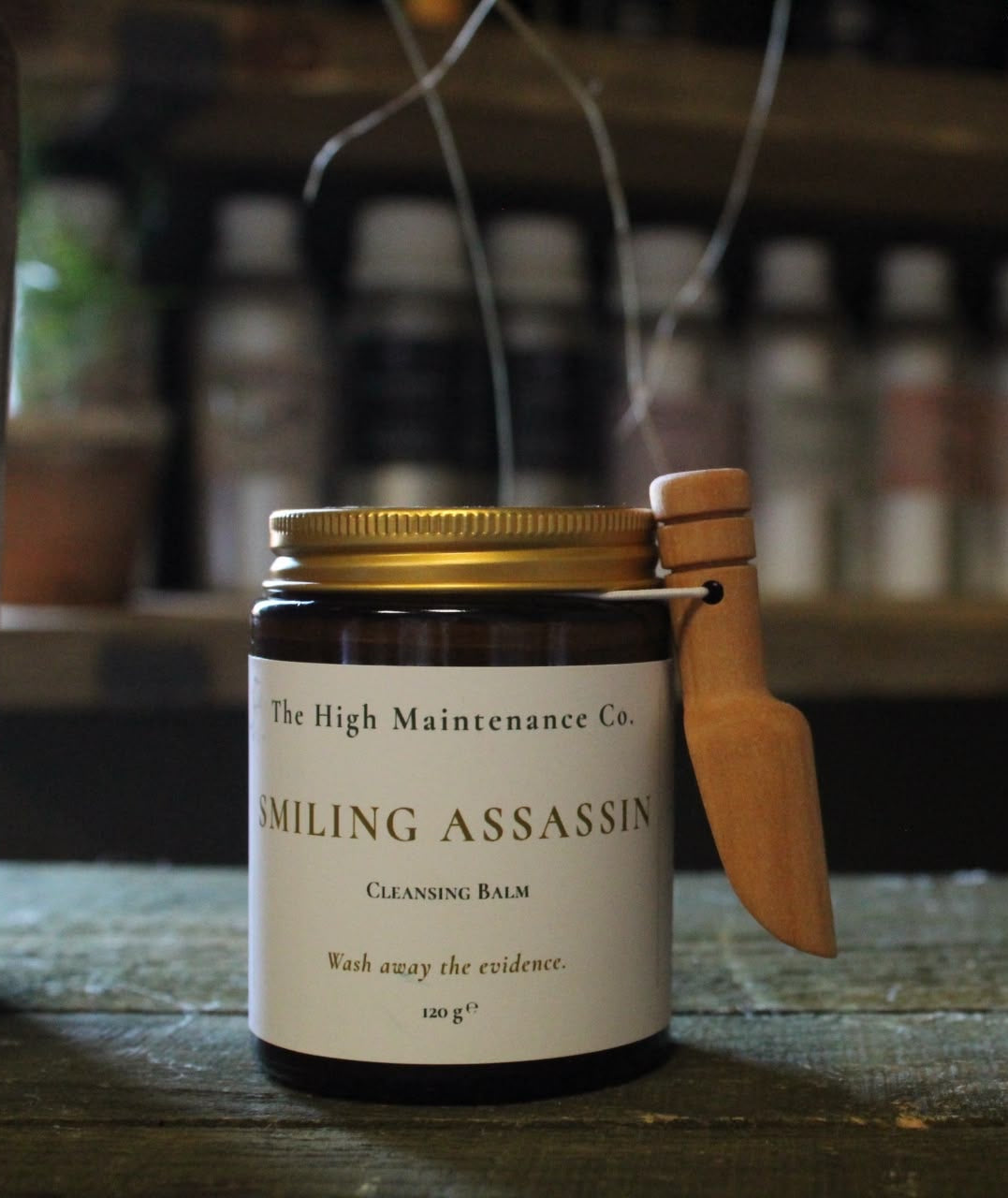 A dark amber glass jar of 'Smiling Assassin' cleansing balm by 'The High Maintenance Co.' sits on a rustic wooden table.