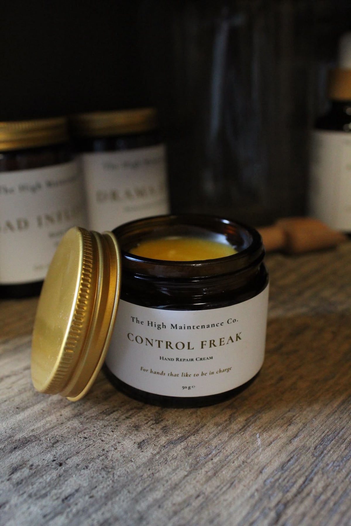 Amber  jar  labeled 'CONTROL FREAK' Hand repair cream by The High Maintenance Co. sits open on a rustic wooden surface, with its gold lid leaning beside it.