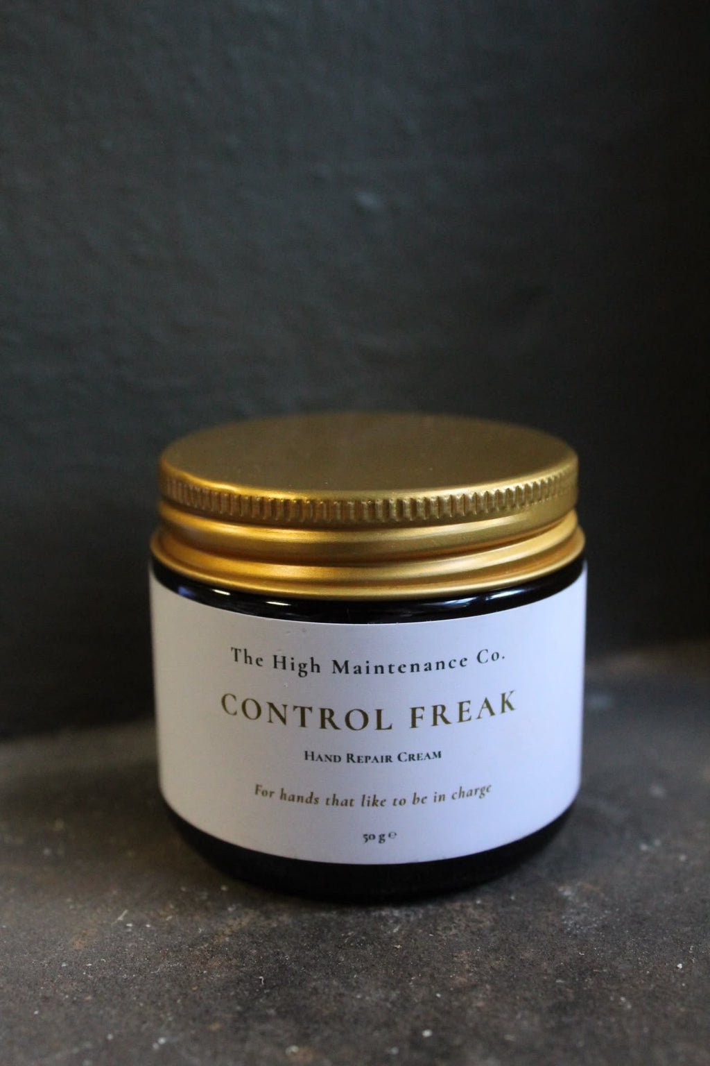 A small dark glass jar with a metallic gold screw-on lid and a white label reading 'The High Maintenance Co. CONTROL FREAK HAND REPAIR CREAM' on a dark textured background.
