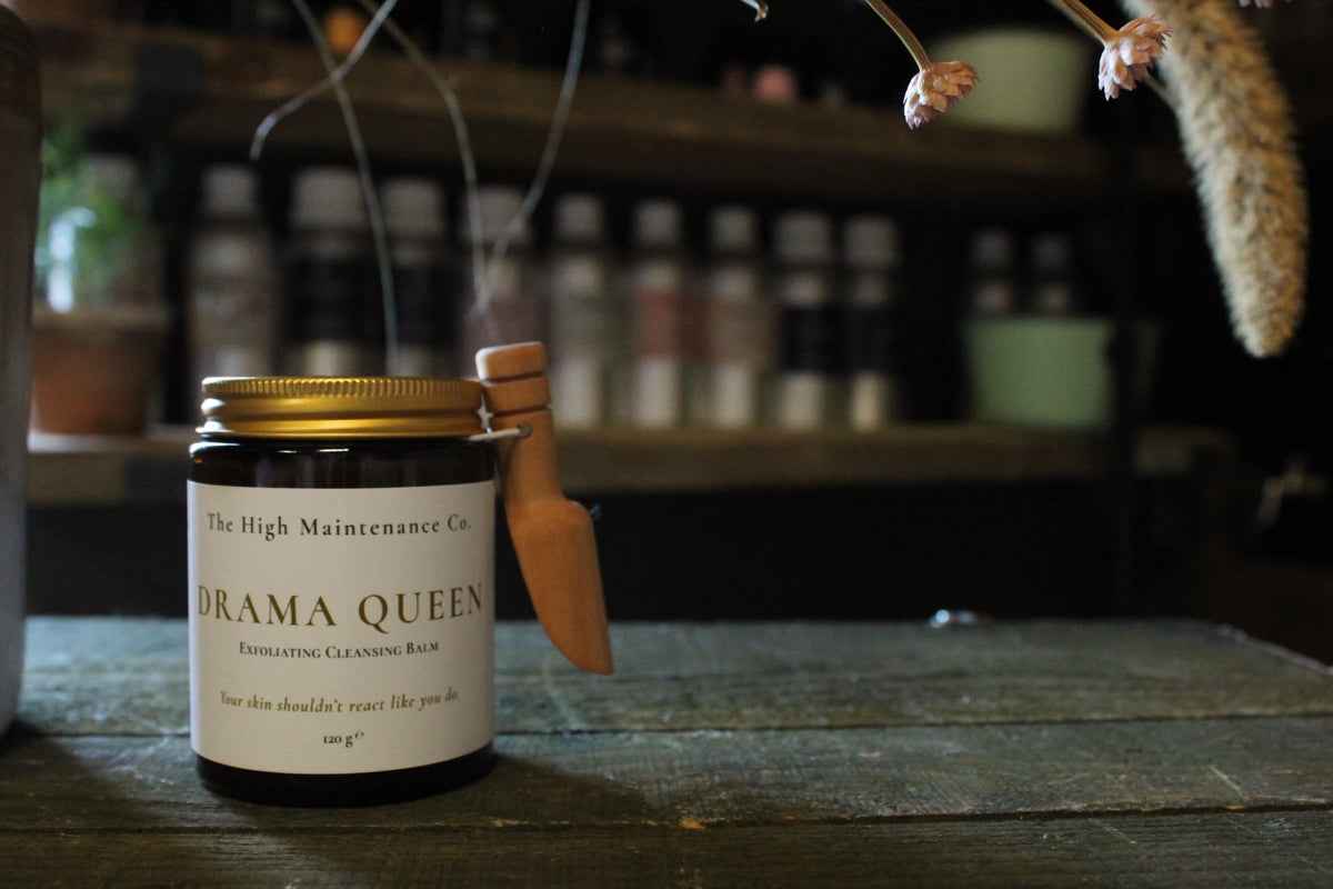 A jar of  exfoliating cleansing balm from The High Maintenance Co. labelled 'Drama Queen' sits on a rustic wooden table with a small wooden scoop resting against the lid.