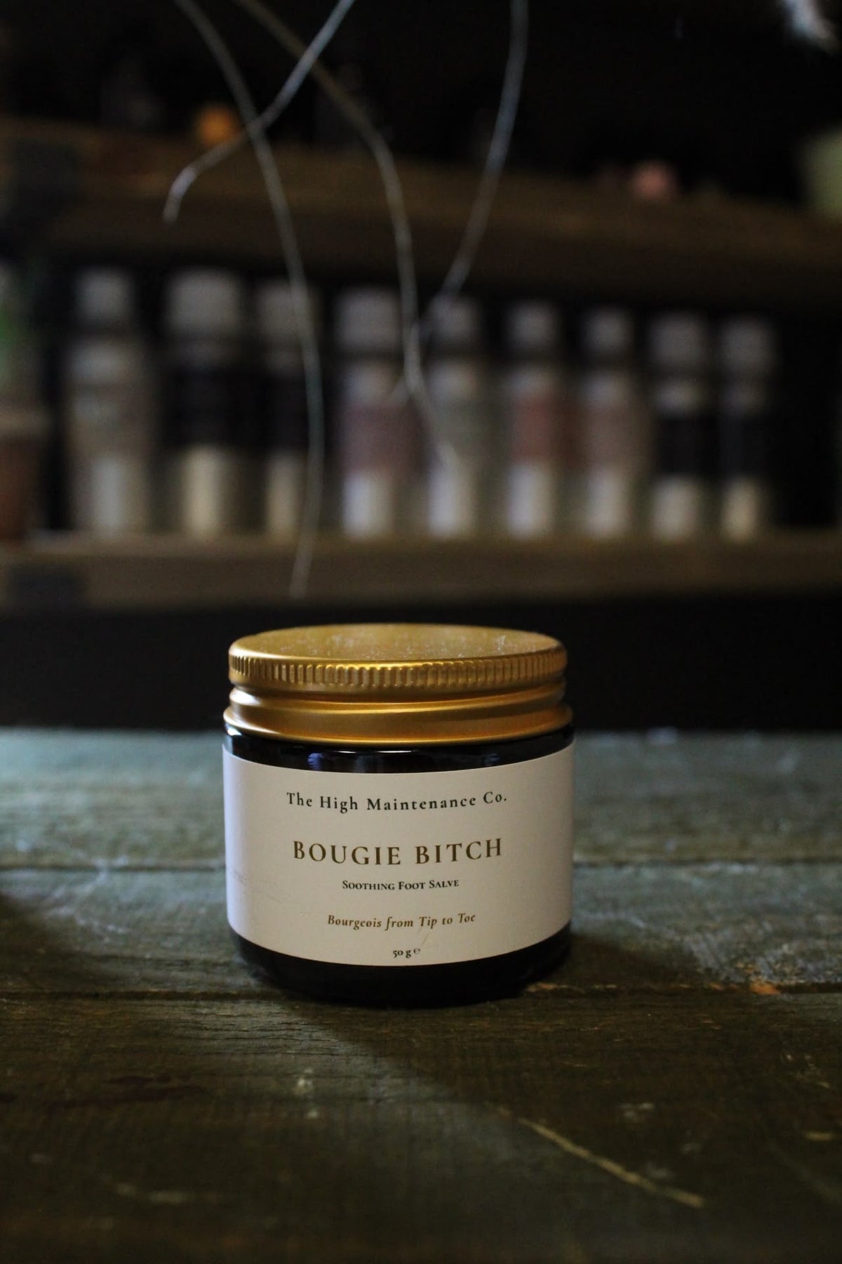 A small amber glass jar with a gold metal lid and a white label reading 'The High Maintenance Co. BOUGIE BITCH' sits on a dark wooden surface with a blurred shop background.
