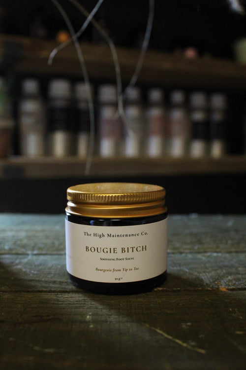 A small amber glass jar with a gold metal lid and a white label reading 'The High Maintenance Co. BOUGIE BITCH' sits on a dark wooden surface with a blurred shop background.