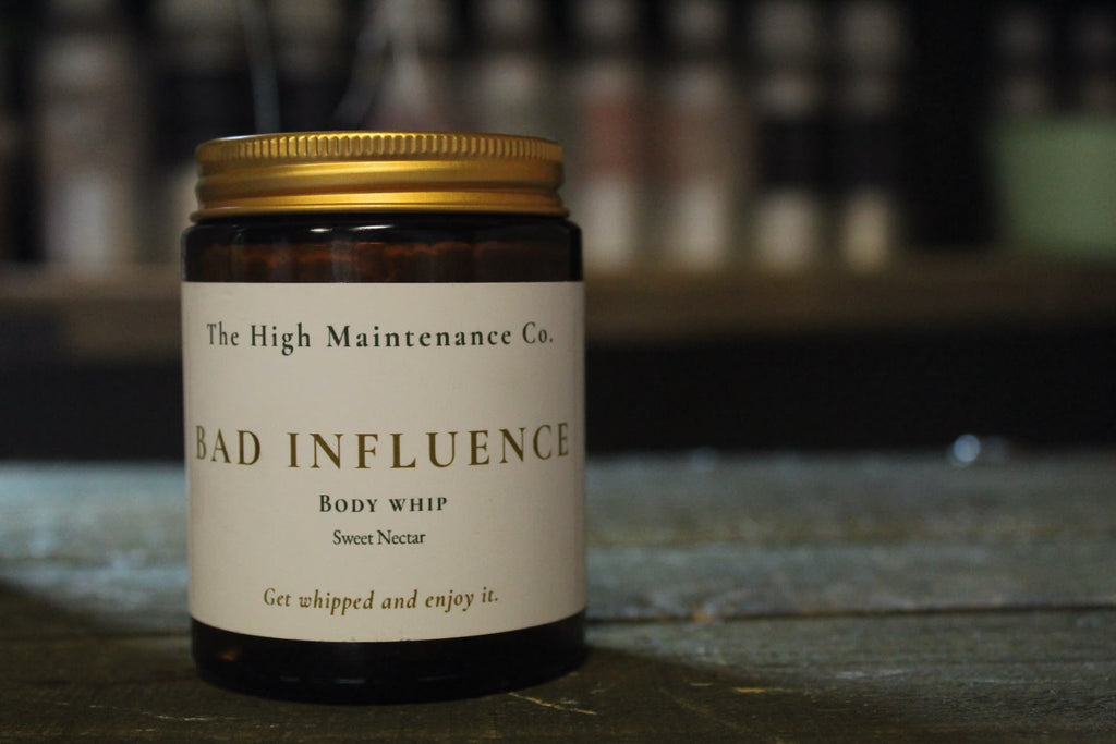 A dark amber glass jar containing honey and vanilla scented body whip with a gold metal lid and a cream-colored label. The label reads 'The High Maintenance Co.