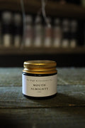 A small glass jar with a white label reading 'MOUTH ALMIGHTY' lip balm and 'The High Maintenance Co.' on the lid, which is gold and sits on a rustic wooden surface with a blurred background.