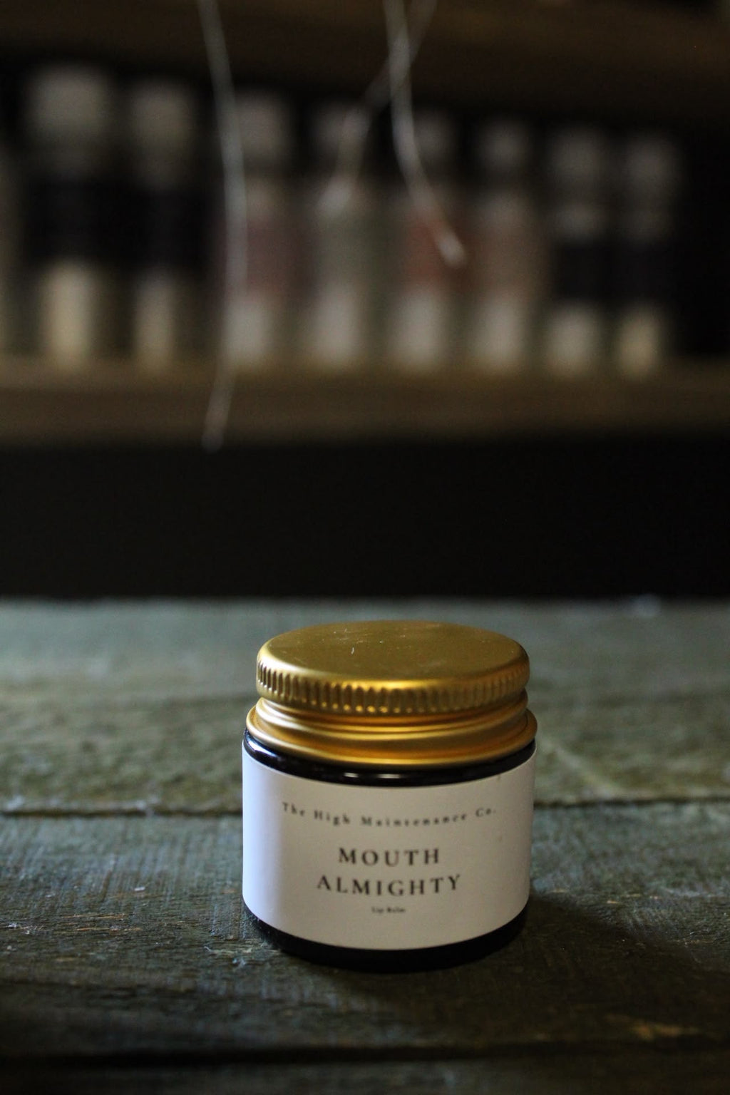 A small glass jar with a gold metal lid and a white label reading 'MOUTH ALMIGHTY' lip balm and 'The High Maintenance Co.' sits on a dark wooden surface with a blurred shelf of similar jars in the background.