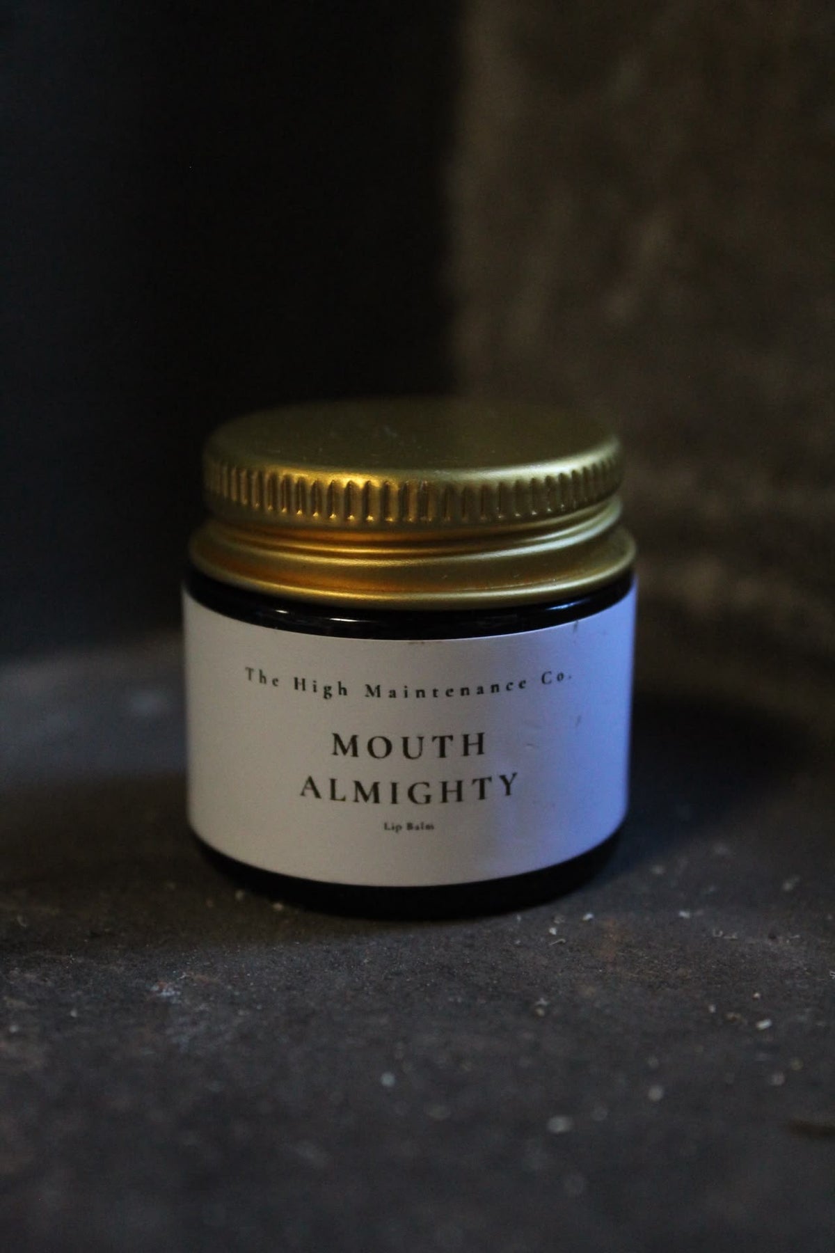 A small amber glass jar with a gold metal screw-top lid and a white label reading 'The High Maintenance Co. MOUTH ALMIGHTY' on a dark textured surface.