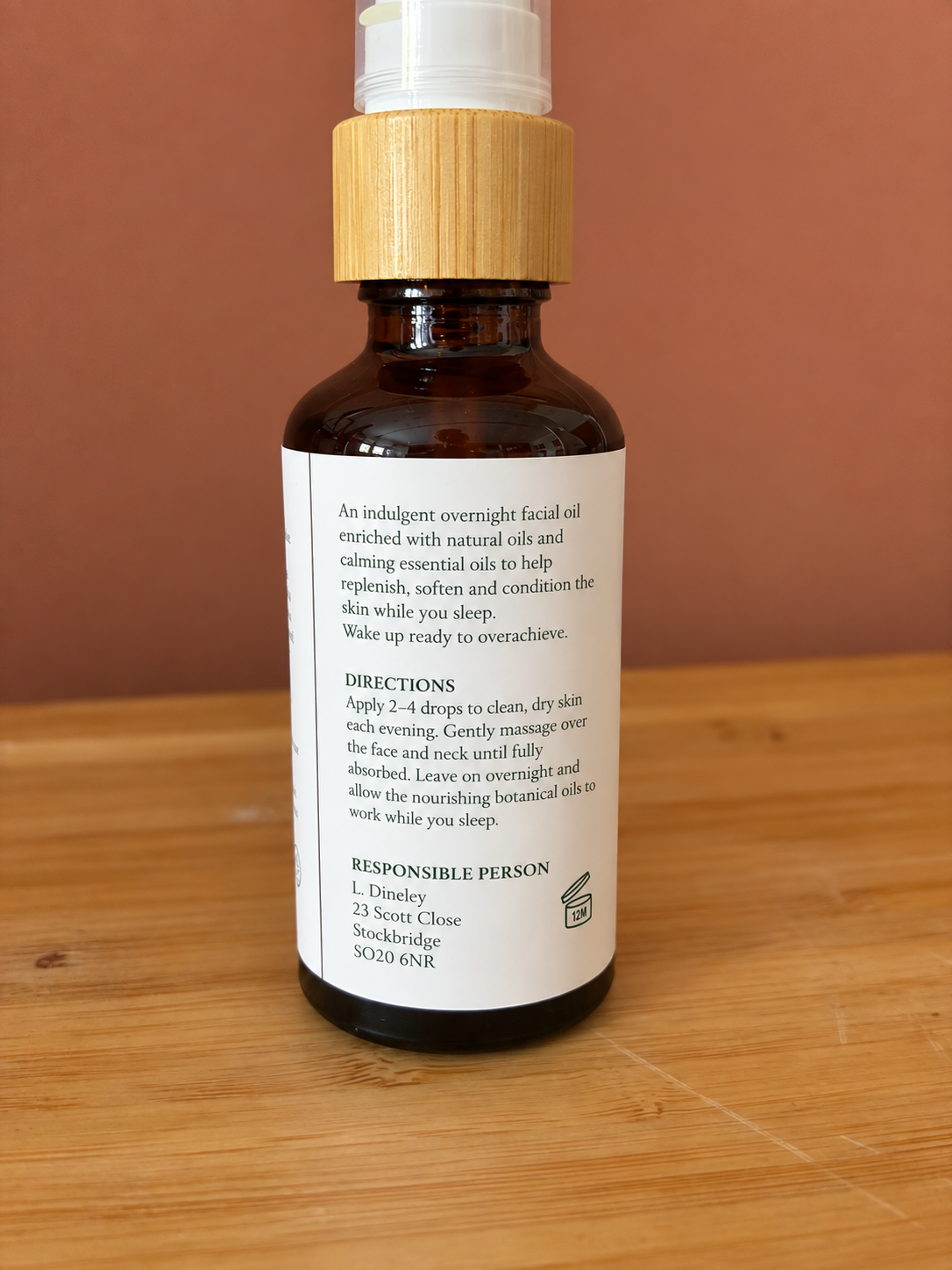 An amber glass facial oil bottle with a light wood pump top and a white pump, standing on a wooden surface against a terracotta background. The front label is turned away, showing the back text of a facial oil product.