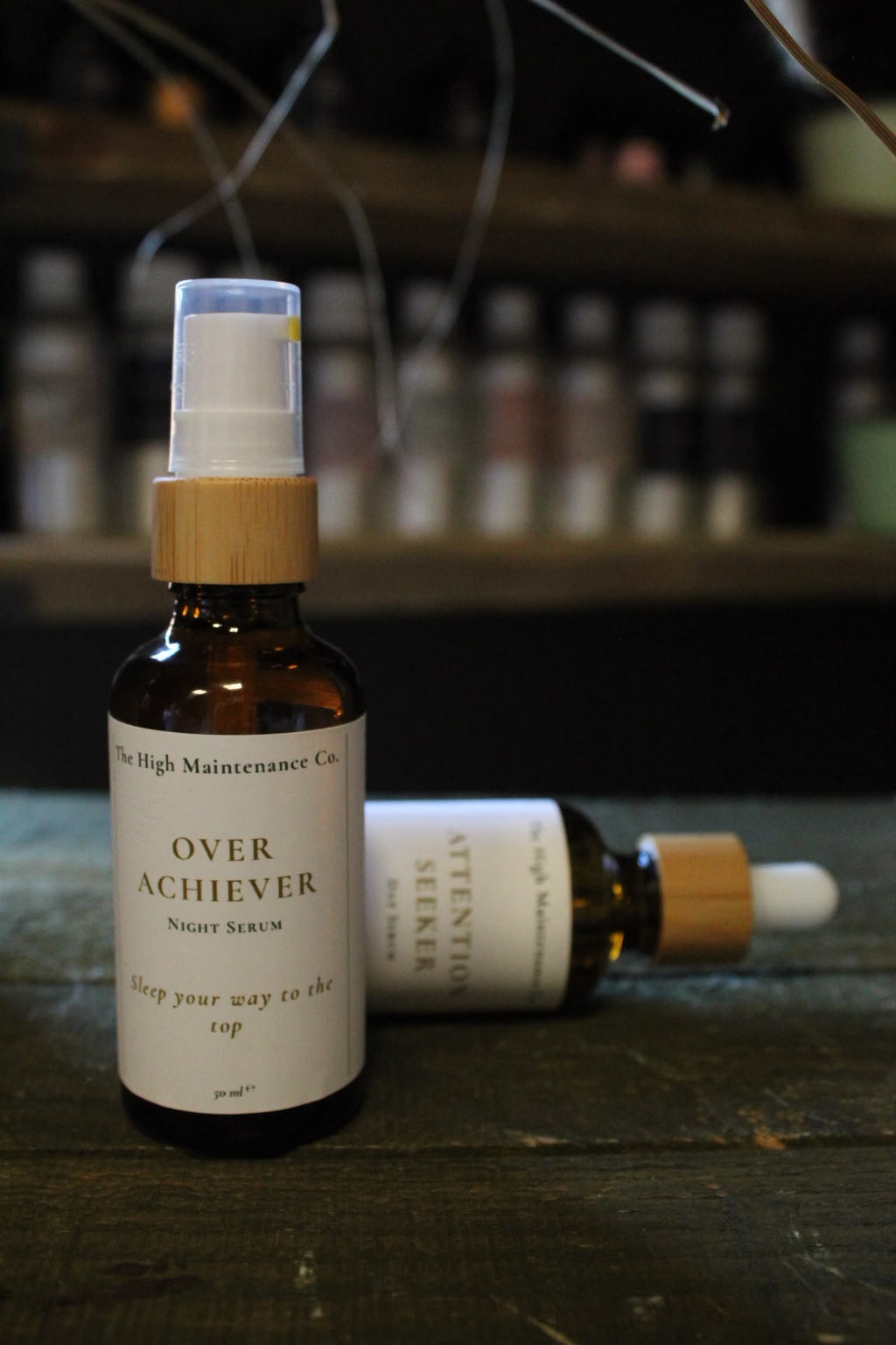 Two amber glass serum bottles on a rustic wooden table, with one standing upright labeled 'OVER ACHIEVER NIGHT SERUM' and one lying down showing 'ATTENTION Seeker Day Serum'.