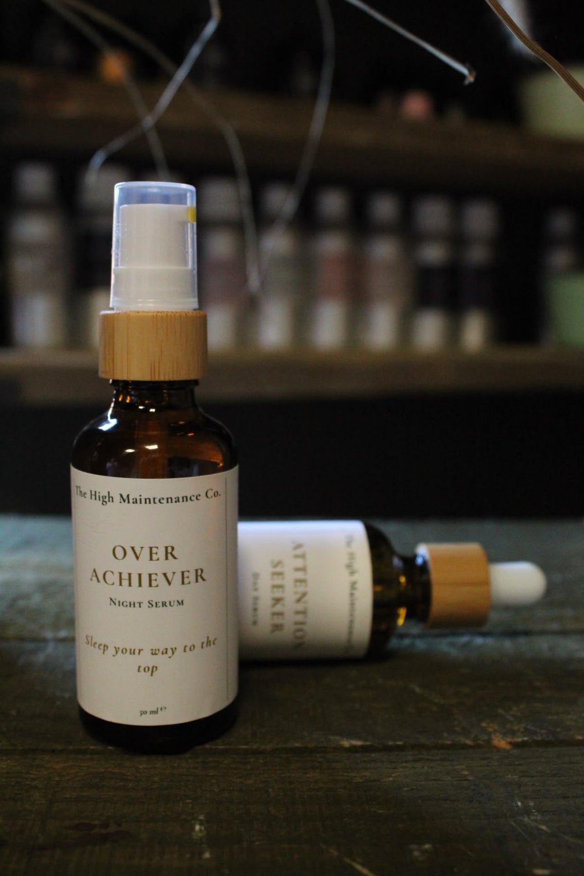 Two amber glass serum bottles on a rustic wooden table, with one standing upright labeled 'OVER ACHIEVER NIGHT SERUM' and one lying down showing 'ATTENTION Seeker Day Serum'.