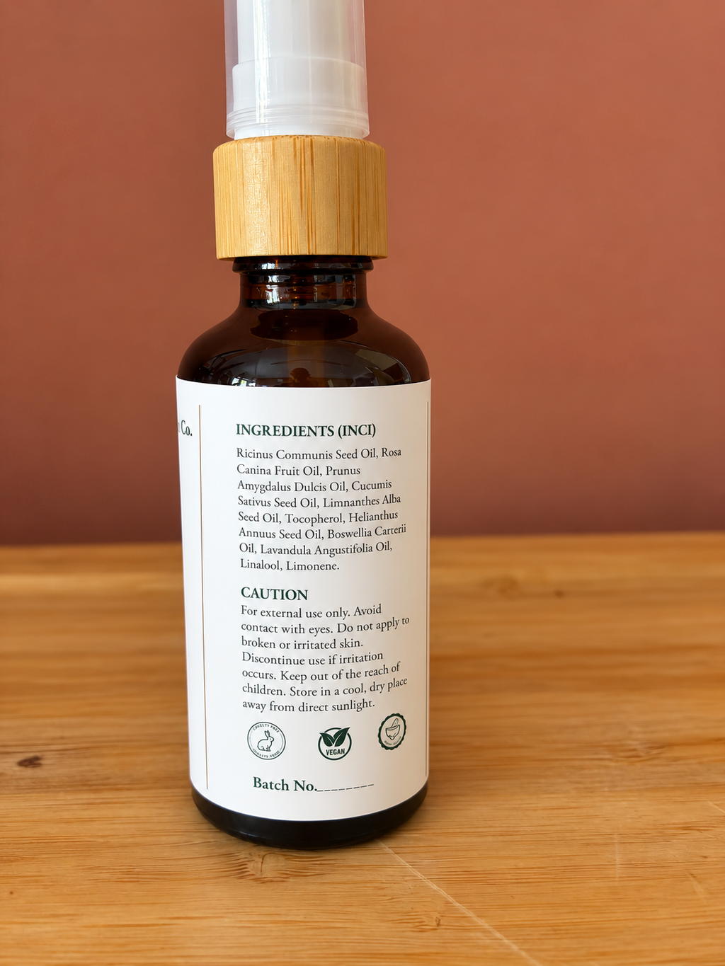 An amber glass bottle of face oil with a light wood cap and white label, standing on a wooden surface against a terracotta background. The back of the label is visible, showing ingredients and caution text.