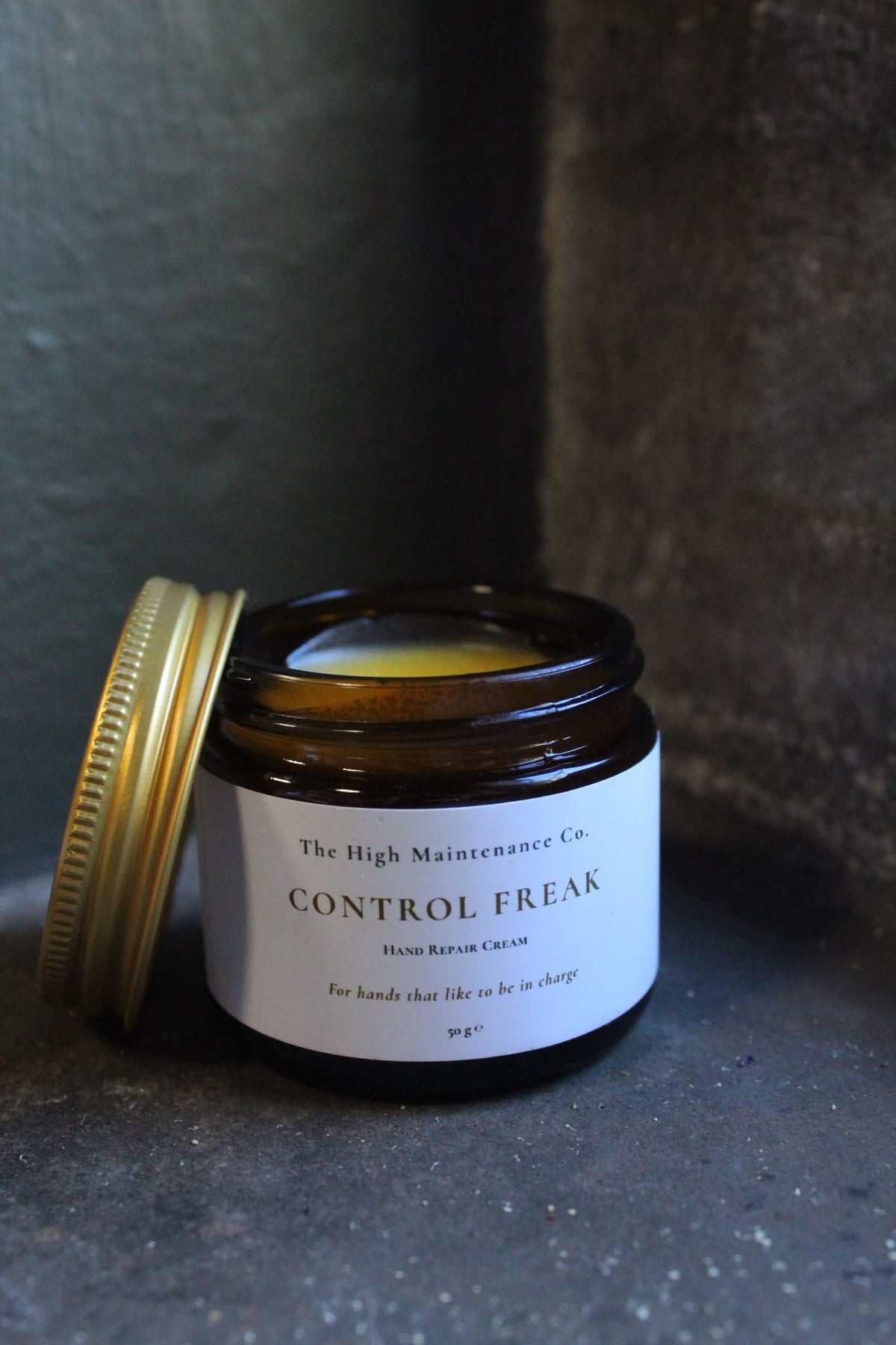 An amber glass jar with its gold metal lid resting beside it on a dark textured surface. The label reads 'The High Maintenance Co. Hand cream