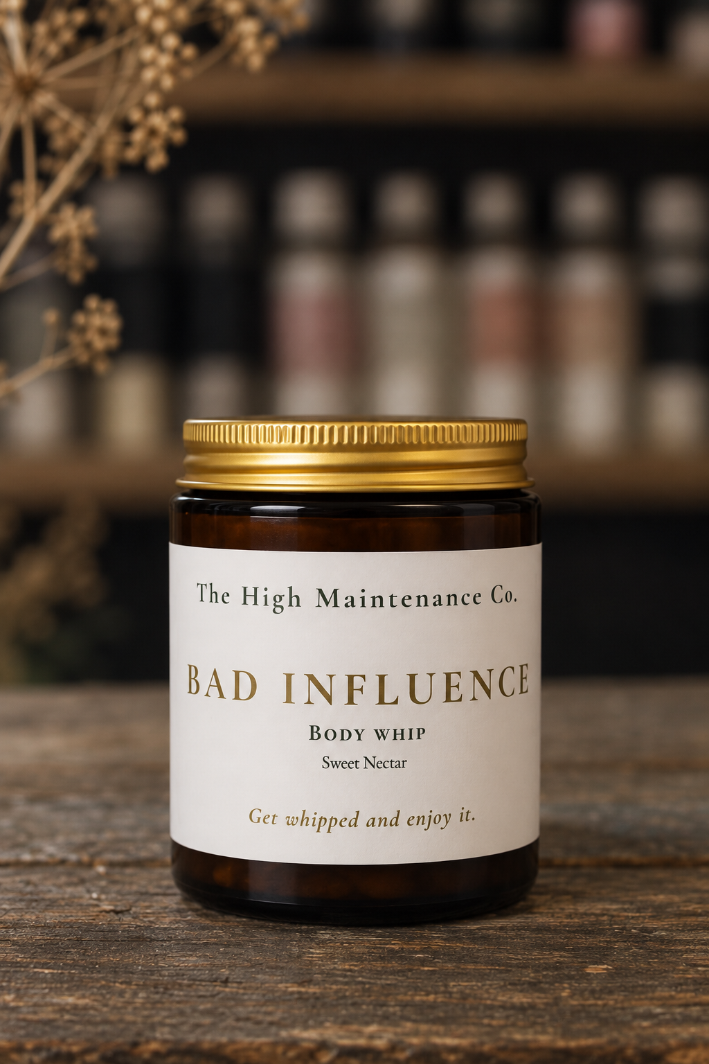 An amber glass jar with a gold metal lid and a white label reading 'The High Maintenance Co. BAD INFLUENCE Body Whip Sweet Nectar' sits on a rustic wooden surface with a blurred shop background.