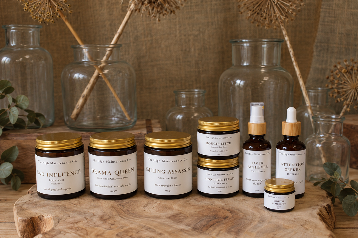 A collection of artisanal skincare and small bottles from The High Maintenance Co, arranged on a rustic wooden slab with glass bottles and dried botanicals in the background.