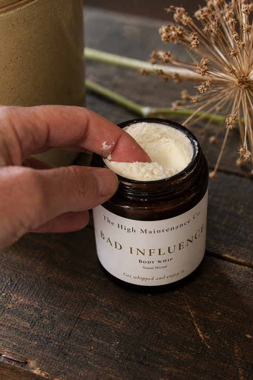 A hand scoops white body whip from an open amber glass jar labeled 'BAD INFLUENCE' by The High Maintenance Co, set on a rustic dark wooden table with dried flowers in the background.